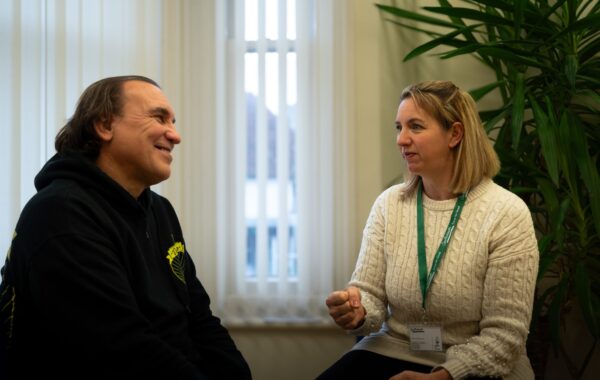 Two people are sitting in conversation in a brightly lit room. On the left, a man with medium-length hair wearing a black hoodie smiles warmly. On the right, a woman with shoulder-length blonde hair wearing a cream cable-knit jumper and a green lanyard gestures as she speaks, appearing engaged. Behind them, a large plant adds a touch of greenery to the space, while vertical blinds cover the window in the background.