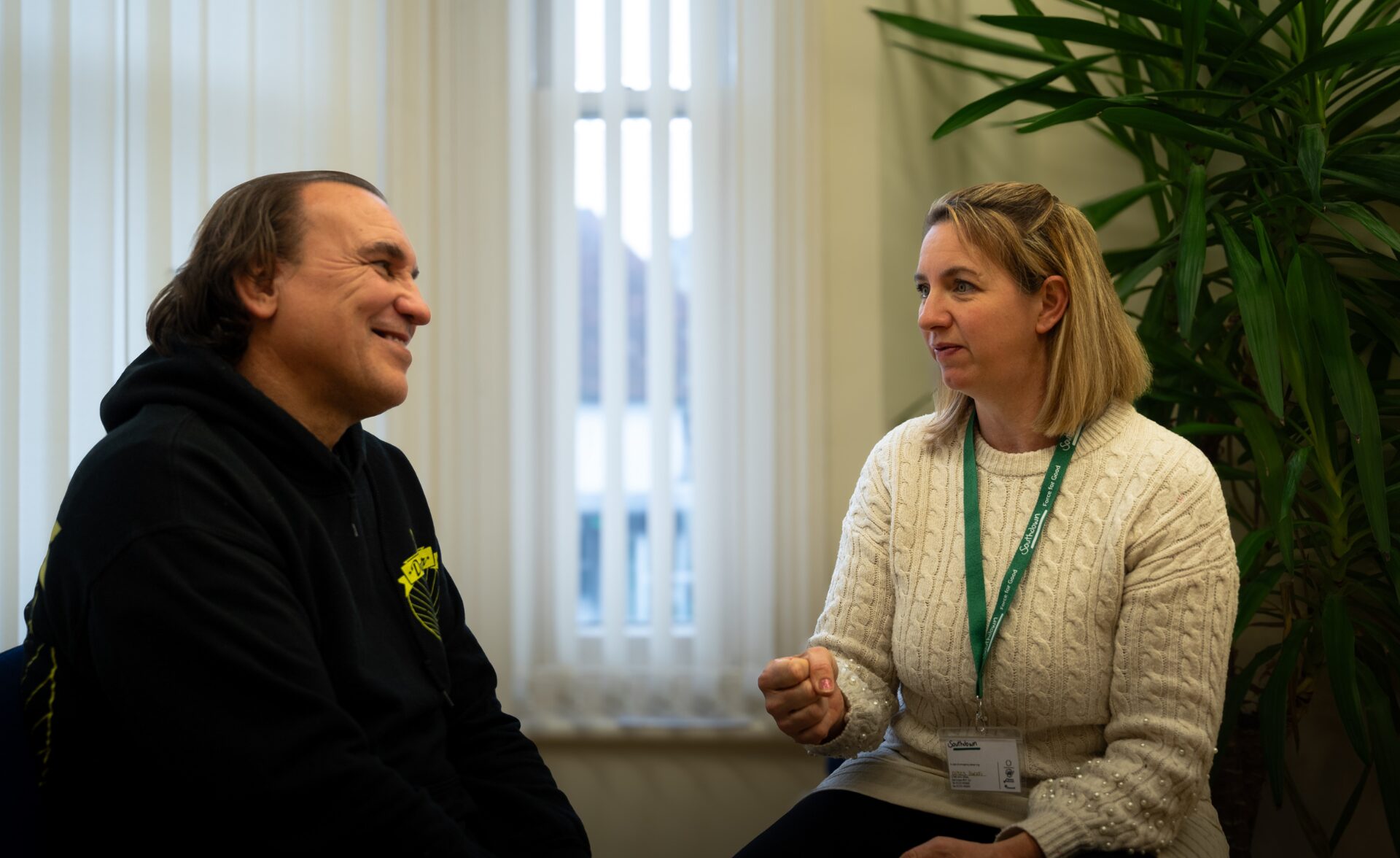 Two people are sitting in conversation in a brightly lit room. On the left, a man with medium-length hair wearing a black hoodie smiles warmly. On the right, a woman with shoulder-length blonde hair wearing a cream cable-knit jumper and a green lanyard gestures as she speaks, appearing engaged. Behind them, a large plant adds a touch of greenery to the space, while vertical blinds cover the window in the background.