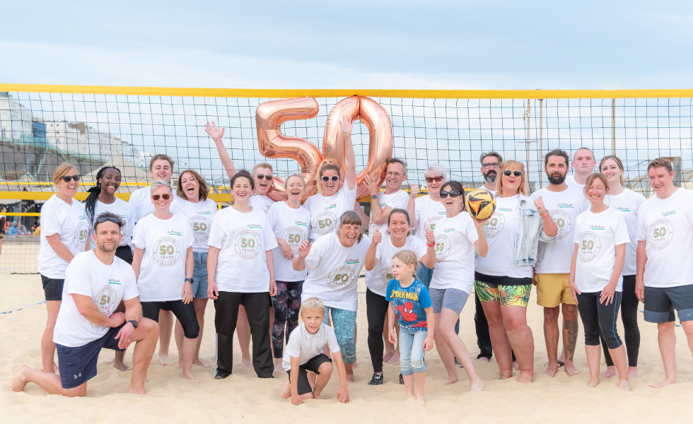 A large group of Southdown colleagues and friends are gathered together celebrating Southdown being around for 50 years. They are on a volley ball court that is sandy near the beach and there is a 50 sign balloon in the middle.