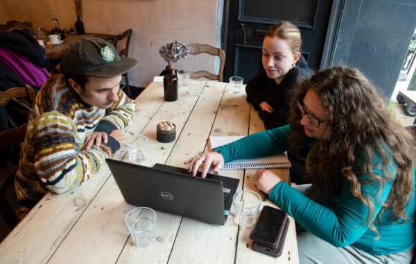 Three people are sat around a wooden table looking at a laptop.