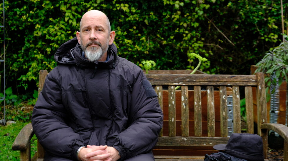 A white man with a smooth head and a grey beard is sitting on a garden bench. He is dressed in black and has a black bag next to him.