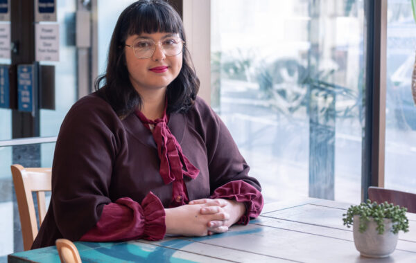 A staff member is sitting down at a table near a window and is looking at the camera.