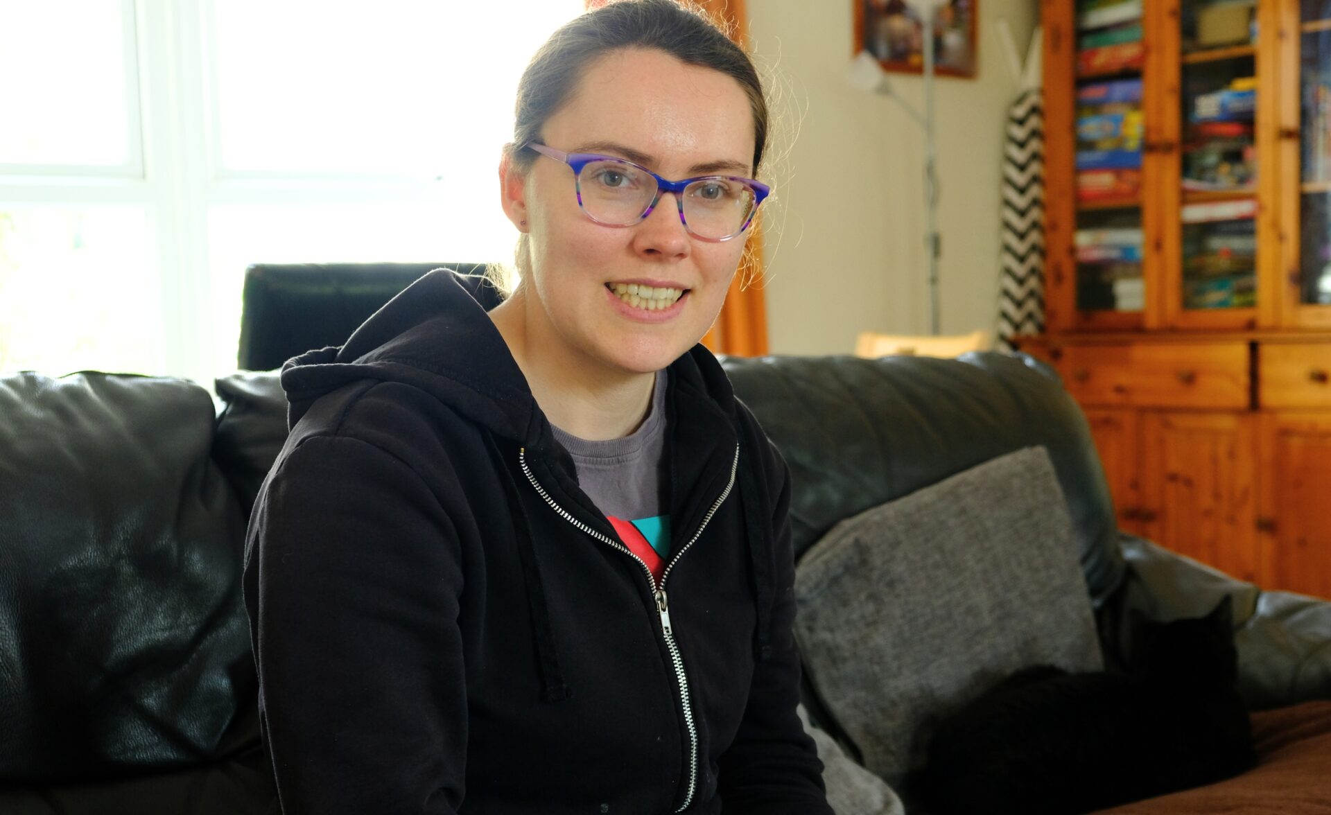 A woman is sitting down on a sofa in a living room. She is smiling whilst looking at the camera.