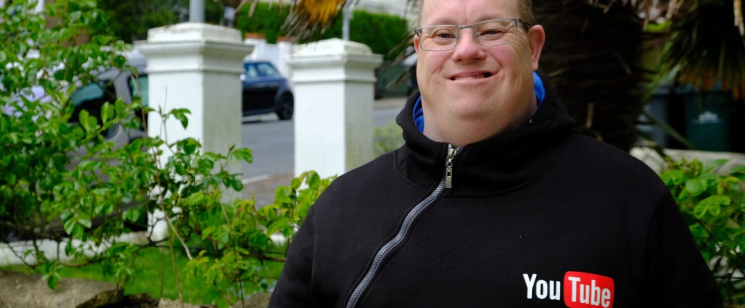 A man is standing outside and is smiling at the camera. He is wearing a black hoodie with a YouTube logo on it.
