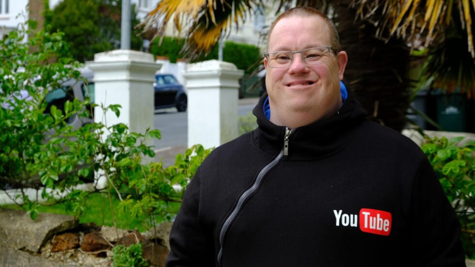 A man is standing outside and is smiling at the camera. He is wearing a black hoodie with a YouTube logo on it.