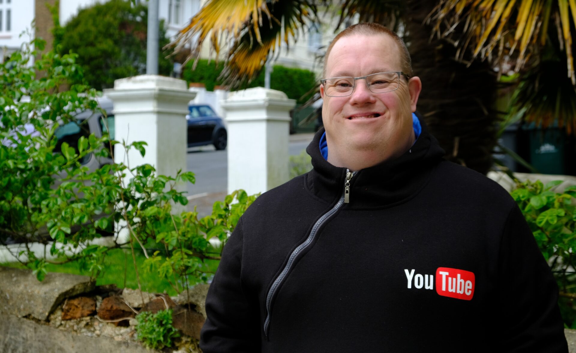 A man is standing outside and is smiling at the camera. He is wearing a black hoodie with a YouTube logo on it.