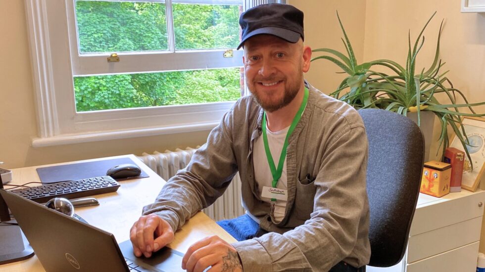 A man is sat at a table inside with a laptop in front of him. Behind him is a window and a plant.