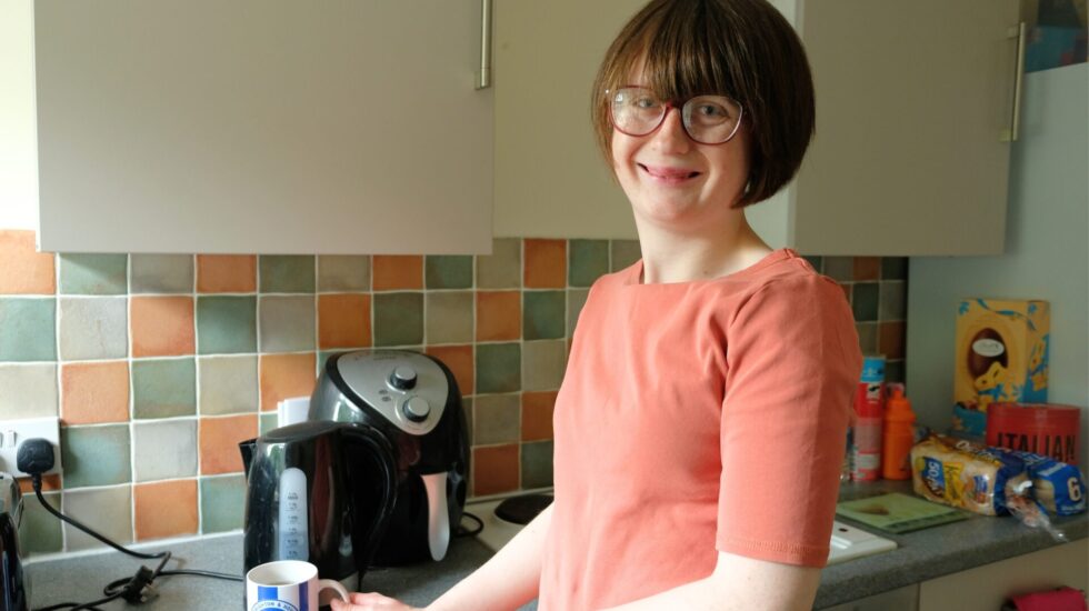 A woman is making a hot drink in the kitchen, she is looking into the camera and smiling.