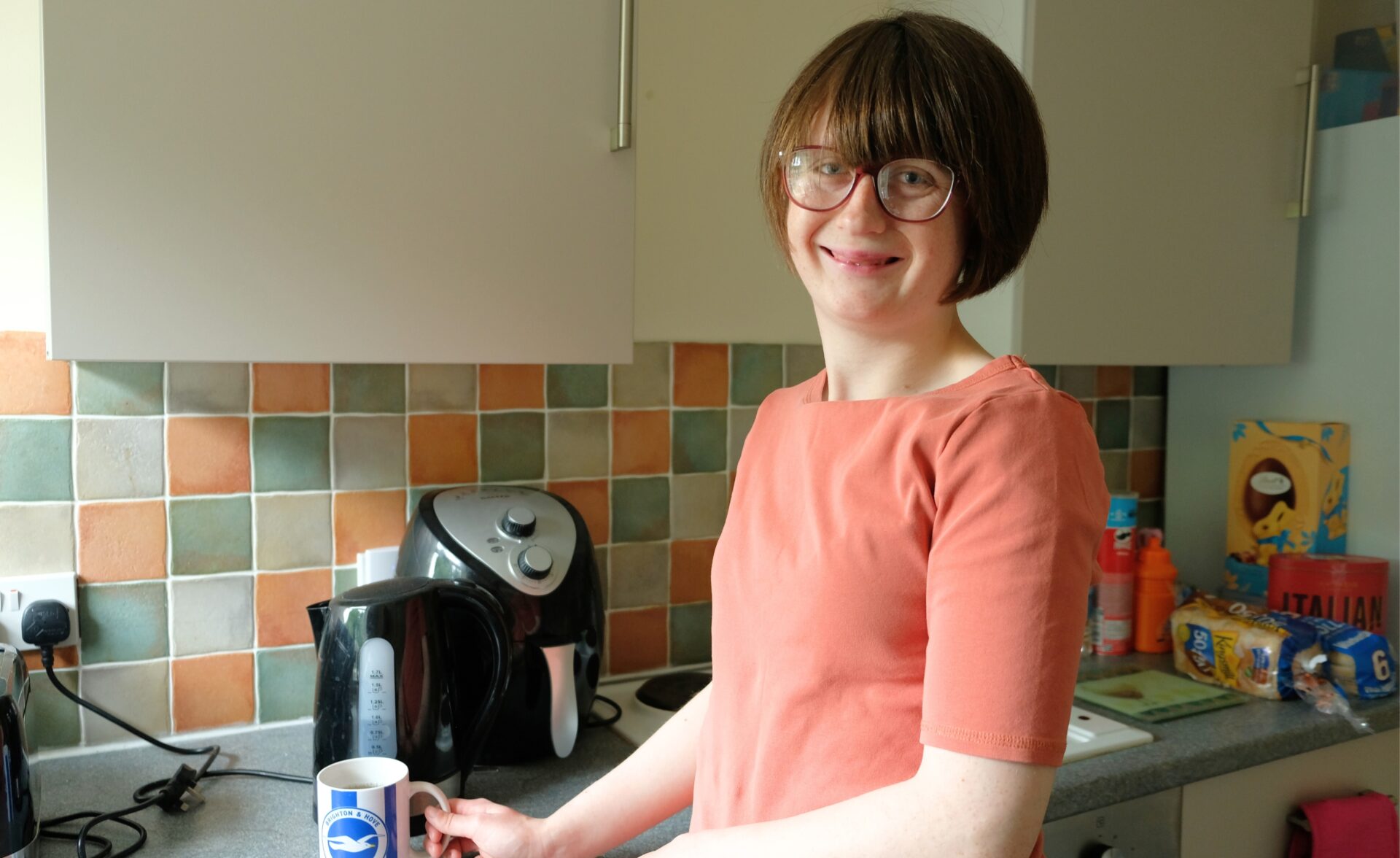 A woman is making a hot drink in the kitchen, she is looking into the camera and smiling.