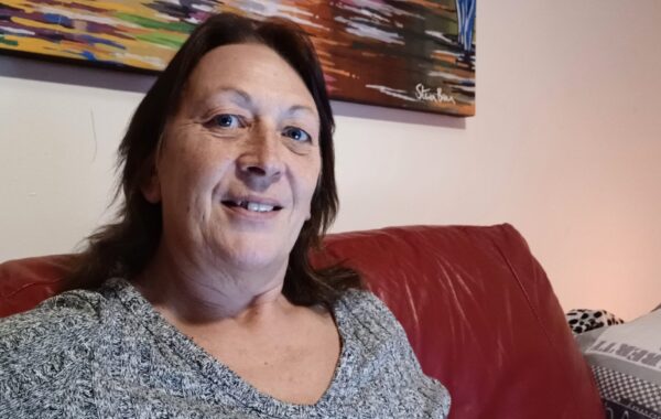 Tracy, Southdown's Housing First Worker wearing a grey dress and looking into the camera with a smiling face, while sitting on a red couch.