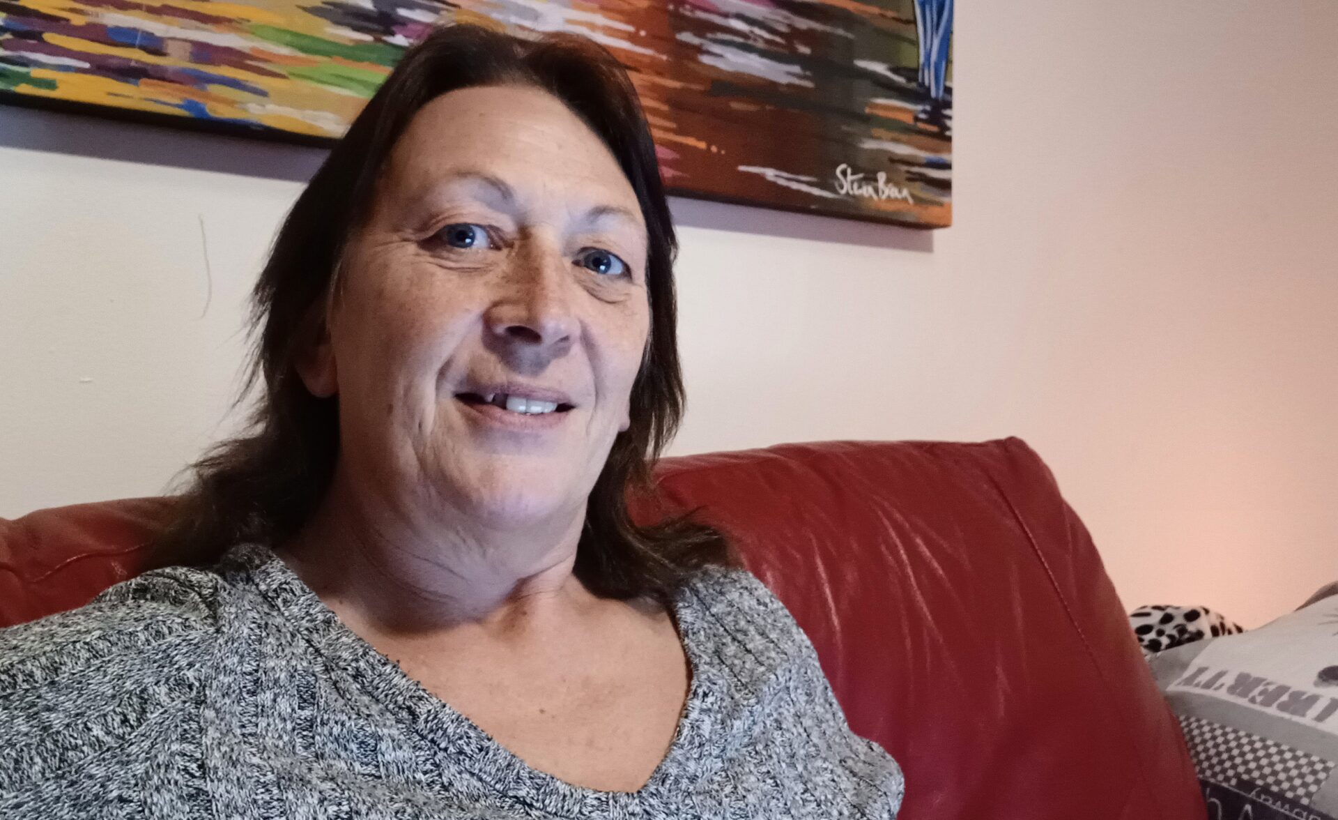 Tracy, Southdown's Housing First Worker wearing a grey dress and looking into the camera with a smiling face, while sitting on a red couch.