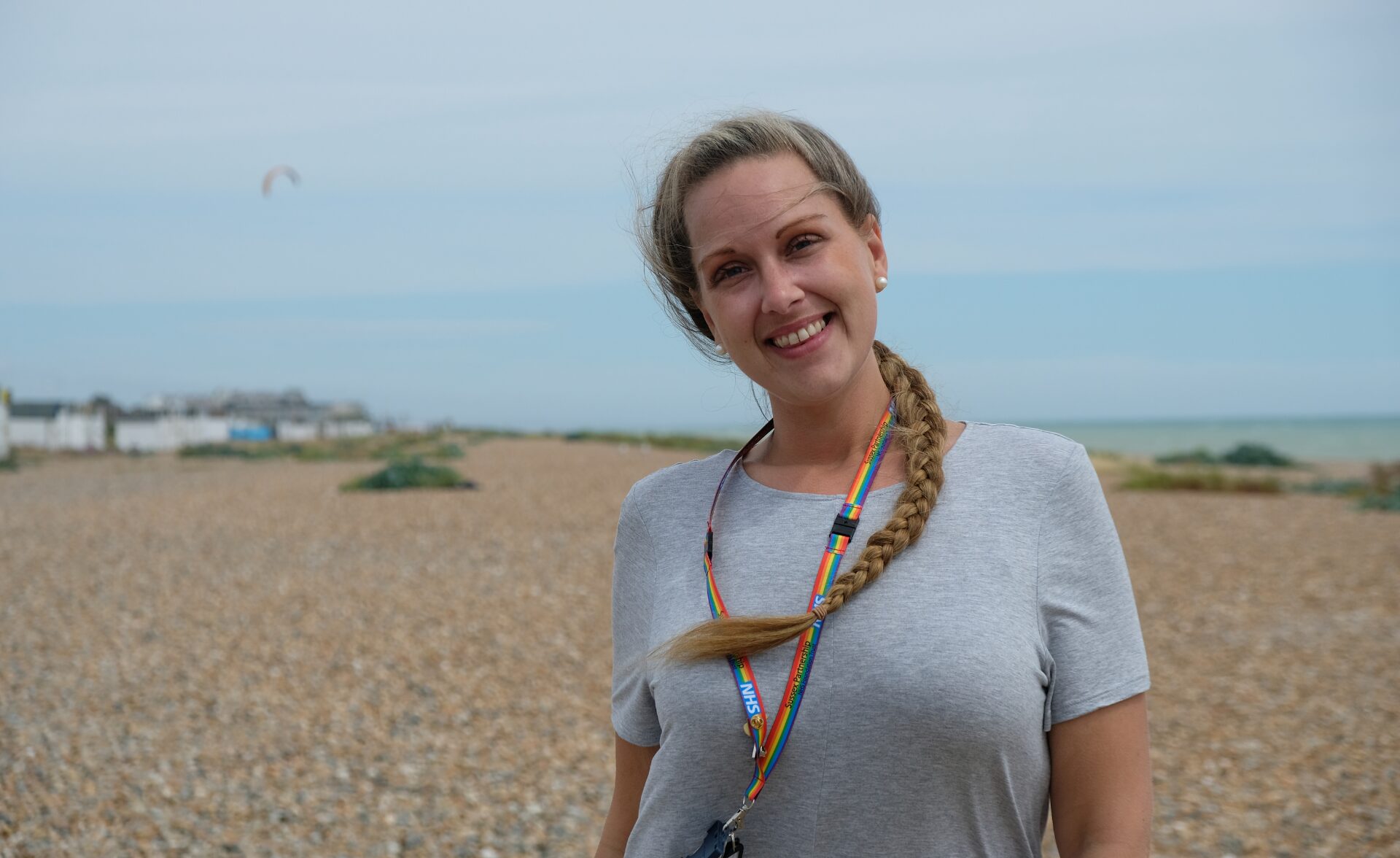 A woman in white tshirt smiles looking at the camera. There is sea in the background, and pebbled beach in sight.