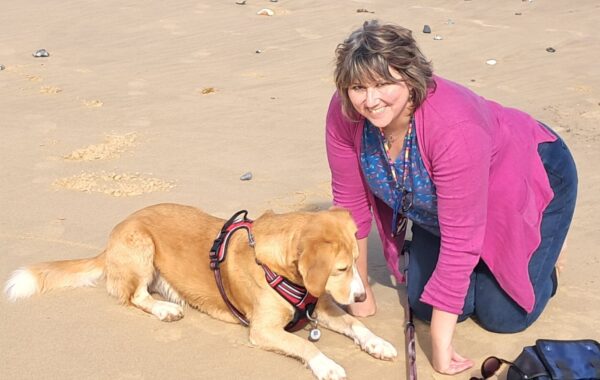 A woman, dressed in pink cardigan and blue tshirt, smilingly looks while she kneels beside her pet dog.