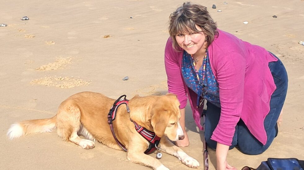 A woman, dressed in pink cardigan and blue tshirt, smilingly looks while she kneels beside her pet dog.