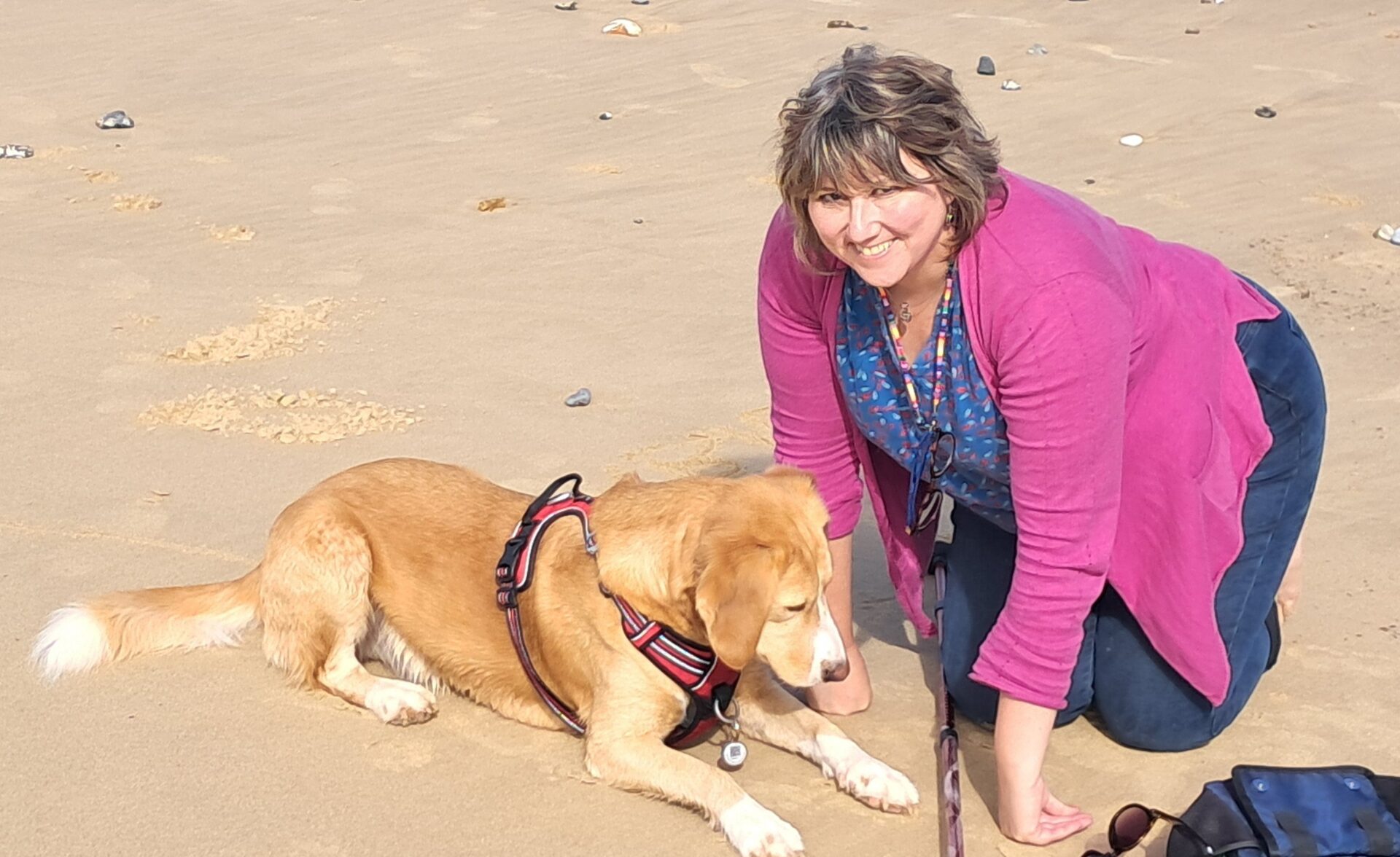 A woman, dressed in pink cardigan and blue tshirt, smilingly looks while she kneels beside her pet dog.