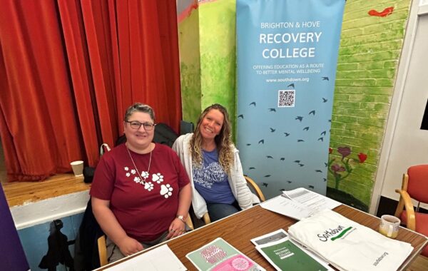 Two women seated at a table with informational materials for the Brighton & Hove Recovery College. Behind them is a banner that reads "Brighton & Hove Recovery College - Offering education as a route to better mental wellbeing" with the website "www.southdown.org" and a QR code. The background features a red curtain and a painted wall with a nature theme.