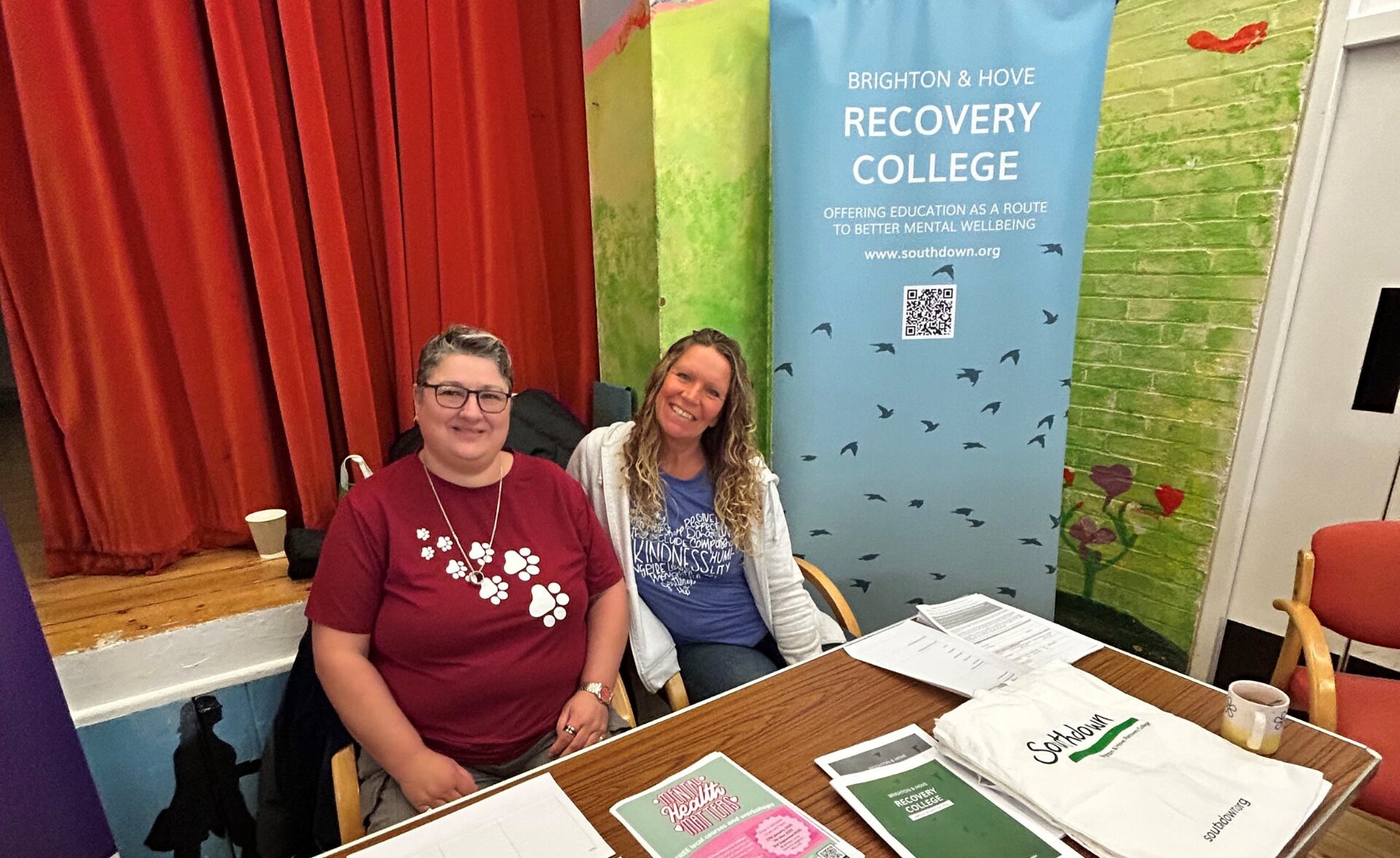 Two women seated at a table with informational materials for the Brighton & Hove Recovery College. Behind them is a banner that reads "Brighton & Hove Recovery College - Offering education as a route to better mental wellbeing" with the website "www.southdown.org" and a QR code. The background features a red curtain and a painted wall with a nature theme.