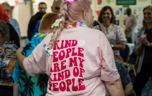 A person with pink hair styled in a braid wearing a pink T-shirt that reads "Kind people are my kind of people" in bold red text with a daisy design. They are participating in a lively Learning Disability Service event.