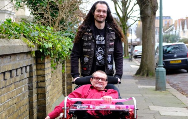 A man with long dark hair, wearing a black heavy metal-themed vest with patches, is pushing a wheelchair along a pavement. The wheelchair user, a client, a man with glasses and a bright red shirt, is smiling. The wheelchair has decorations, including stickers with political messages. They are outdoors on a tree-lined street with brick houses and parked cars in the background. They are featured in government's 'Made with Care' campaign.