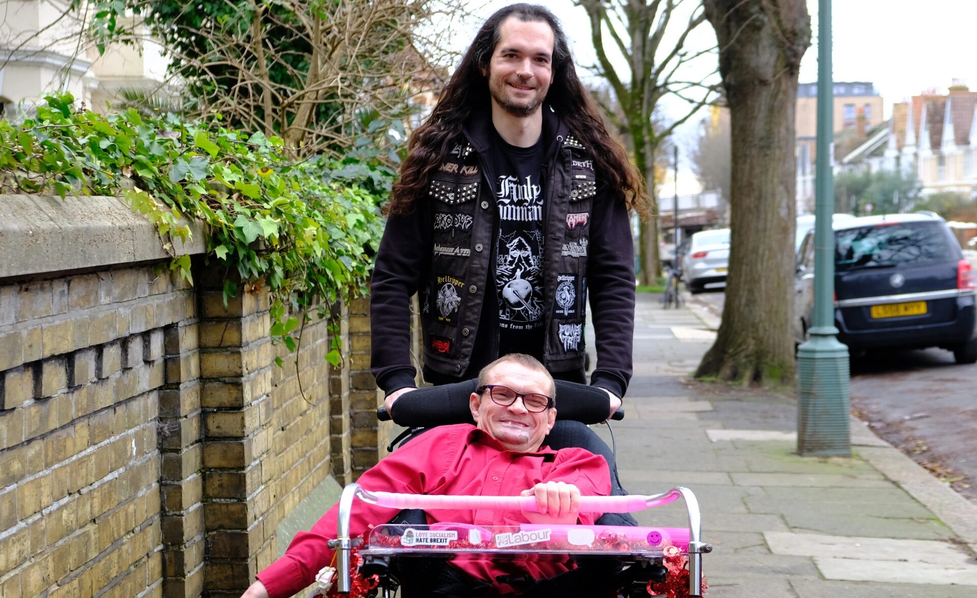 A man with long dark hair, wearing a black heavy metal-themed vest with patches, is pushing a wheelchair along a pavement. The wheelchair user, a client, a man with glasses and a bright red shirt, is smiling. The wheelchair has decorations, including stickers with political messages. They are outdoors on a tree-lined street with brick houses and parked cars in the background. They are featured in government's 'Made with Care' campaign.