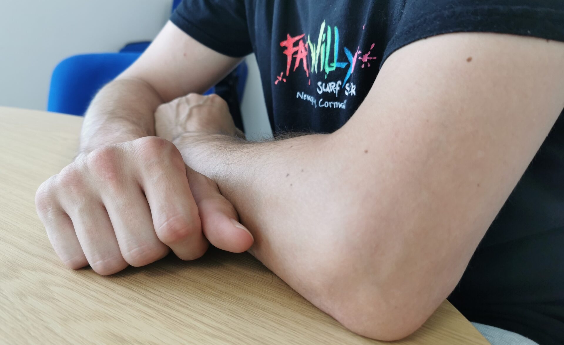 A close-up photograph of a person’s arms resting on a light wooden table. The individual is wearing a black T-shirt with a colourful "Family Surf" logo. The person is a client of the Pathways Home service, a Housing Support programme in West Sussex. The image conveys a sense of stability and support, reflecting the service's goal of helping individuals build independence through responsive housing support.